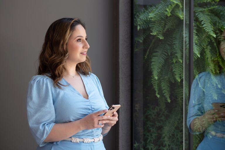 Uma mulher em uma sala com o celular na mão, olhando para o lado de fora e sorrindo levemente.